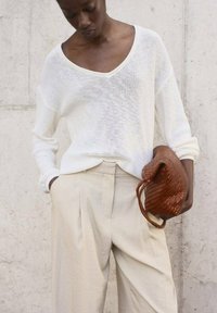 Pull en maille lâche blanc avec un col en V et des manches longues, associé à un pantalon taille haute beige clair ; tient un petit sac tressé marron.