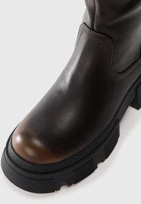 Botte en cuir marron foncé avec semelle en caoutchouc noir épais et talon carré, finition lisse avec coutures ton sur ton et bout arrondi.
