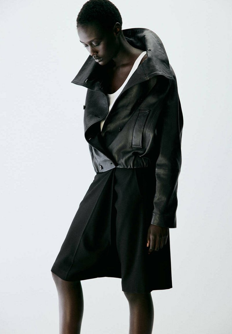Femme aux cheveux courts portant une veste en cuir noire oversize par-dessus un haut blanc et un short noir à jambes larges sur un fond uni.