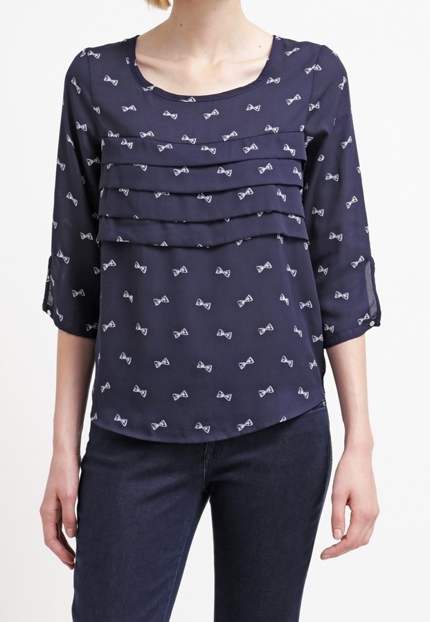 Blouse bleu marine en tissu léger, avec trois volants en couches sur la poitrine et un motif de petits nœuds blancs. Manches trois-quarts.