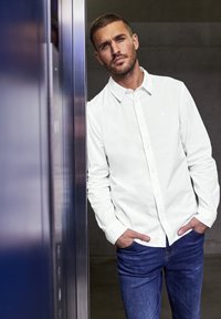 Camicia bianca con colletto, maniche lunghe e un logo discreto; abbinata a jeans in denim blu scuro. Texture liscia, design casual.