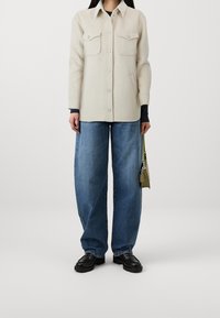 Abrigo de botón beige claro con dos bolsillos en el pecho, combinado con jeans azules holgados y zapatos negros. Bolso verde en la mano derecha.