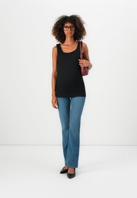 Kvinne med krøllete hår som har på seg briller, svart tanktop, blå jeans, svarte hæler og holder en burgunder skulderveske mot en enkel hvit bakgrunn.