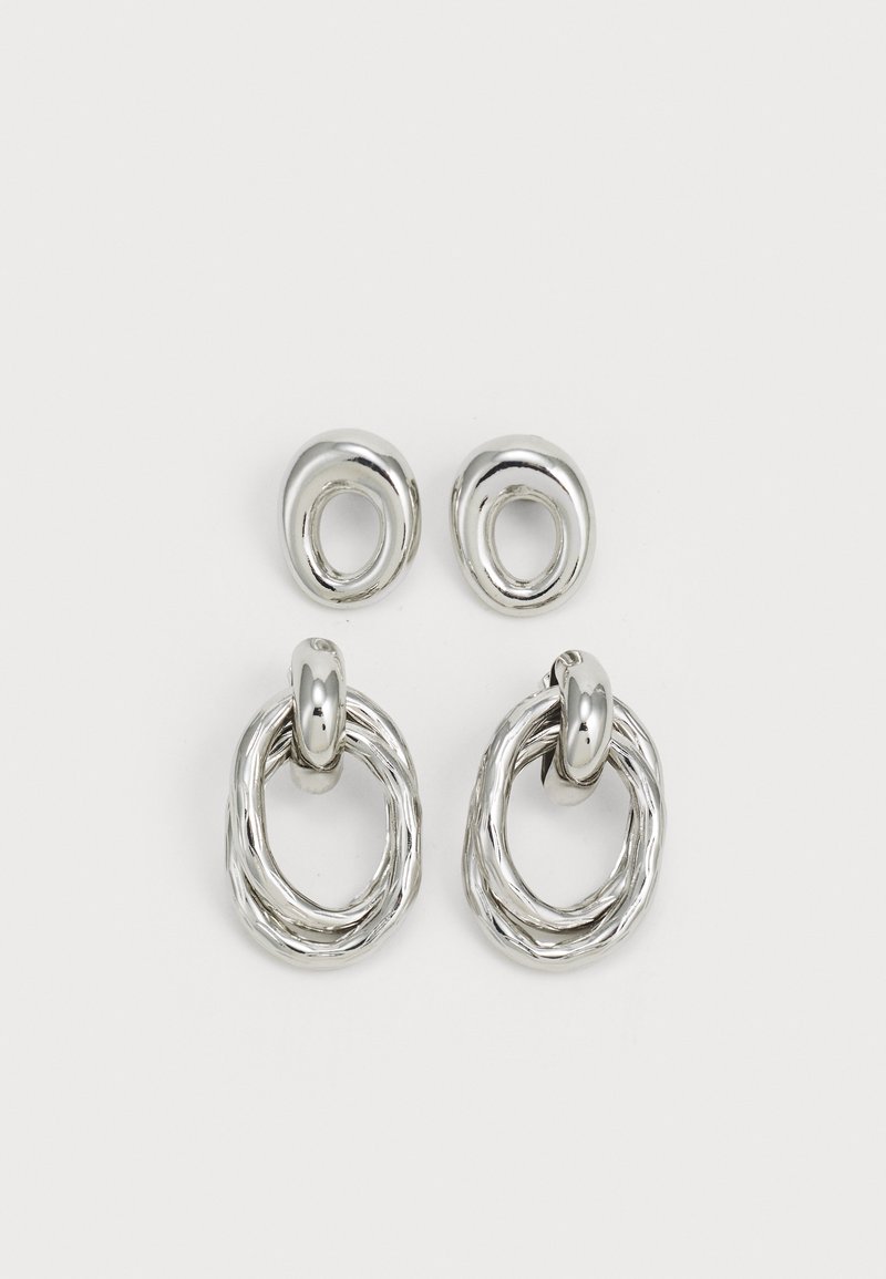 Pendientes de metal tono plateado que presentan dos diseños: un aro circular retorcido y un botón circular liso. La superficie reflectante realza la elegancia.