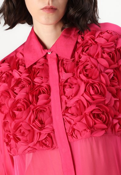 Blouse rose vif avec un design floral texturé composé de tissus superposés, dotée d'un col pointu et d'une patte de boutonnage centrale.