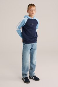 Sötétkék kapucnis pulóver világoskék ujjal és felirattal, világoskék, koptatott farmernadrággal és fekete cipővel, fehér csíkokkal.