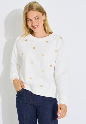 Mujer con cabello rubio vistiendo una sudadera blanca con corazones dorados y vaqueros azules, sonriendo con una mano en el bolsillo.