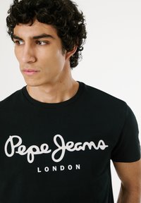 Mladý muž s kučeravými tmavými vlasmi, oblečený v čiernom tričku s bielym nápisom „Pepe Jeans London“ na prednej strane, pozerá sa doľava.
