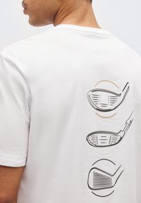 Άνδρας που φοράει λευκό μπλουζάκι με τρία μαύρα περιγράμματα κεφαλών γκολφ τυπωμένα κάθετα στην πλάτη.