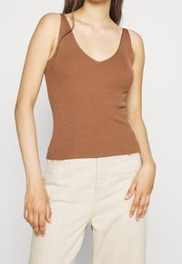 Femme portant un haut marron en tricot côtelé sans manches et un pantalon beige clair taille haute, debout les mains détendues le long du corps.