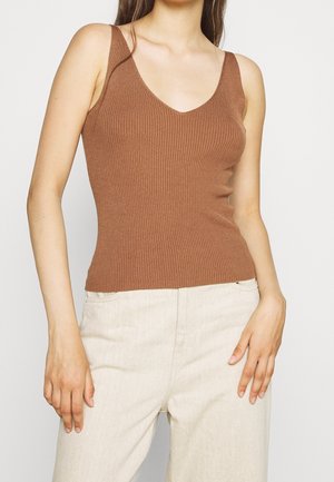 Femme portant un haut marron en tricot côtelé sans manches et un pantalon beige clair taille haute, debout les mains détendues le long du corps.