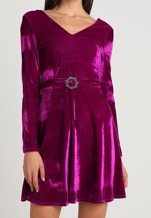 Robe longue en velours magenta profond avec manches longues, décolleté en V, jupe évasée et boucle ronde décorative à la taille.