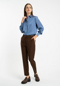 Jasnoniebieska satynowa bluzka z detalami guziczków, w połączeniu z brązowymi dopasowanymi spodniami z boczną kieszenią i brązowymi butami wsuwanymi z grubą podeszwą.