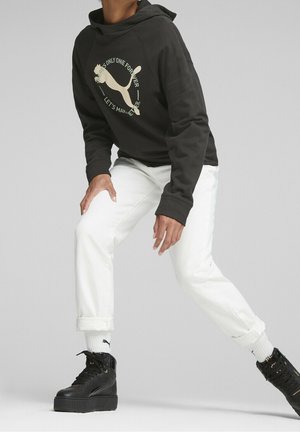 Persona che indossa una felpa nera con cappuccio con il logo Puma, pantaloni bianchi risvoltati, calzini bianchi Puma e sneakers nere con zeppa in posa di stretching.