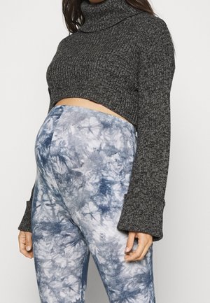 Osoba w ciąży ubrana w ciemnoszary krótki golf i niebiesko-białe legginsy ciążowe typu tie-dye, stojąca na jednolitym tle.
