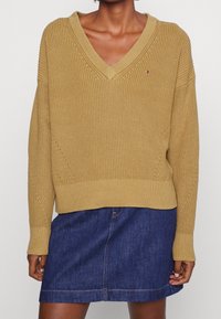 Senfgelbes Strickpullover mit V-Ausschnitt, gerippten Bündchen und Saum, getragen über einem dunklen Jeansrock. Verfügt über eine feine Textur und subtile Akzentdetails.