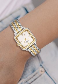 Ruutuline hõbe- ja kuldne kell, valge diali, kuldsete aktsentide ja kahetoonilise metallkettega, kus on ristkülikukujulised lingid.