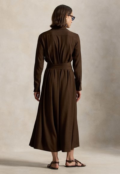 Γυναίκα με κοντά καστανά μαλλιά, φοράει σκούρο καφέ φόρεμα με μακριά μανίκια και ζώνη, καθώς και καφέ σανδάλια, στέκεται με την πλάτη προς τα εμπρός σε μπεζ φόντο.