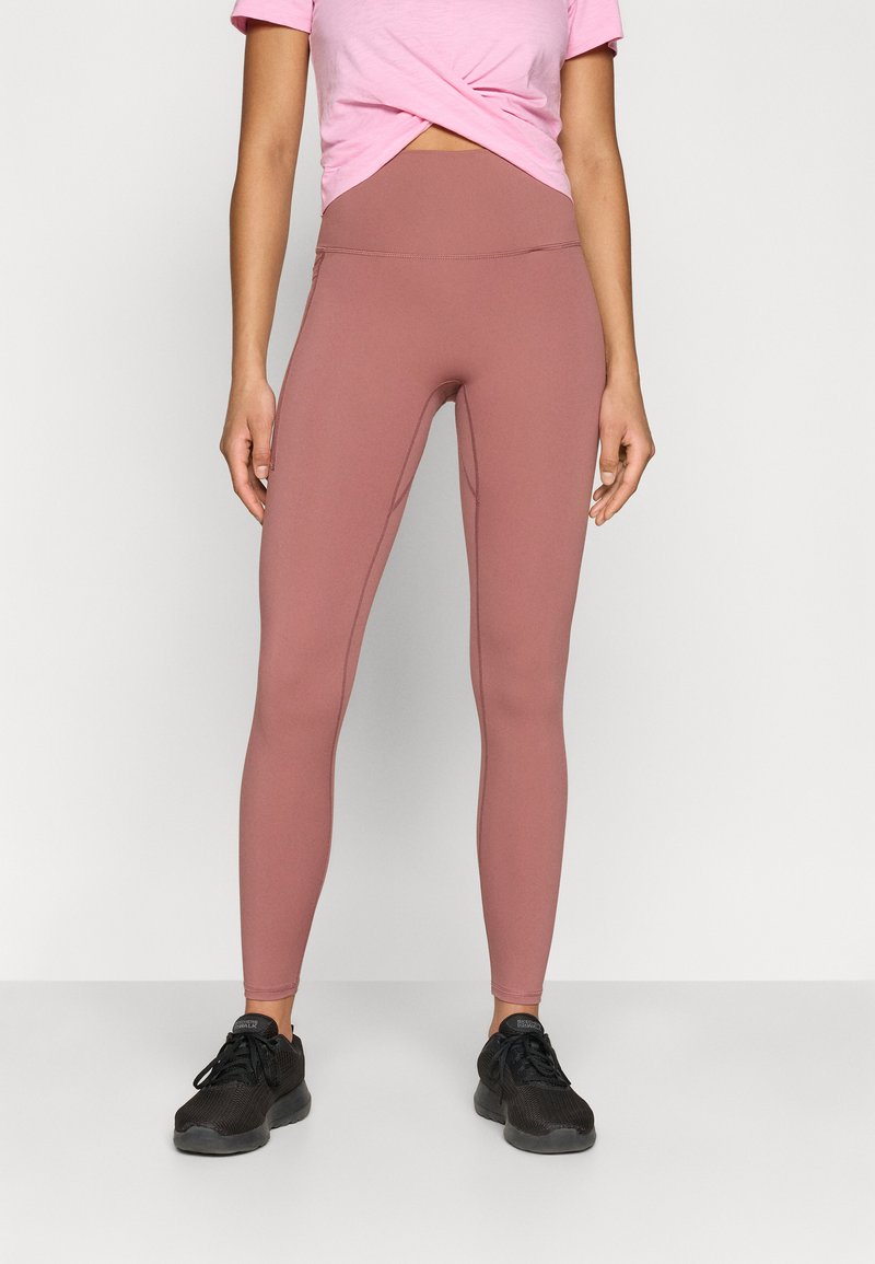 Ροζ κοντομάνικη μπλούζα με στριμμένο μπροστινό μέρος, συνδυασμένη με μεσαίας μέσης, στενά ροζ κολάν σε αποχρώσεις σκόνης. Το μοντέλο φοράει μαύρα αθλητικά παπούτσια.