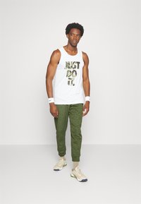 Camisola branca de alças com texto "FAZ ISSO" em padrão de camuflagem, combinada com calças de treino texturizadas verdes e tênis atléticos neutros. Pulseiras brancas.