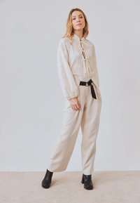 Blusa de encaje crema con cuello alto y cierre de lazo, combinada con pantalones beige de cintura alta y botas negras de tacón cuadrado.