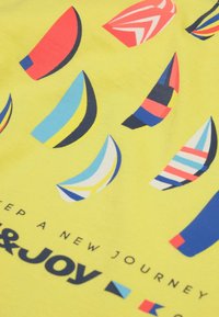Tissu jaune avec des formes abstraites colorées de voiliers et texte partiel indiquant « Commence un nouveau voyage » et « JOIE » en lettres grasses.
