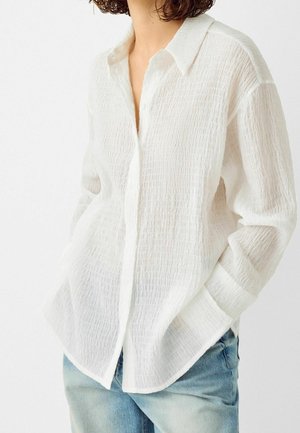 Personne portant une chemise blanche texturée à boutons avec manches longues et un jean en denim bleu clair, mains dans les poches, devant un fond uni.