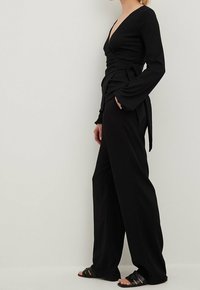 Black wrap top with long sleeves, V-neckline, and side tie, paired with loose black trousers and flat black sandals. Smooth fabric texture.
