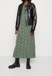Schwarze Lederjacke im Crop-Stil über einem grünen Maxikleid mit wellenförmigem schwarzen Muster. Das Model trägt schwarze Knöchelstiefel, die kontrastierende Texturen zur Schau stellen.