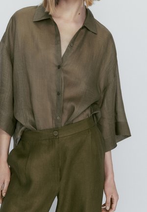 Personne portant une chemise ample transparente olive à boutons avec des manches trois-quarts et un pantalon taille haute assorti sur un fond uni.