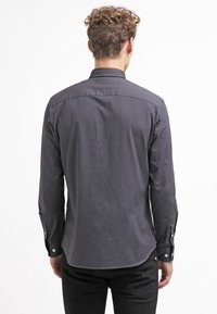 Camicia a maniche lunghe grigio carboncino con colletto a bottone, texture liscia e orlo arrotondato, dotata di polsini con bottoni e spallina sul retro.