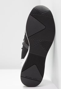 Svarta sneakers med gummisula, texturerad tygöverdel och vit mellansula med Michael Kors-logotyp. Horisontella och diagonala mönster i sulan.