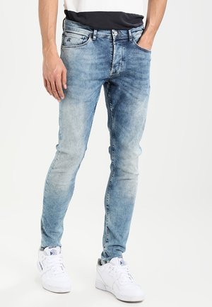 Jeans de mezclilla azul claro con un corte Slim, que presentan un lavado desgastado, ligeras arrugas y un diseño de cinco bolsillos. Llevados con zapatillas blancas.