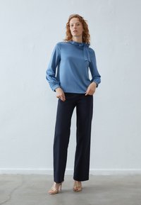 Világoskék selyemblúz, nyakkendővel a gallérnál, hosszú ujjak, és tengerészkék, bő szárú nadrág. A modell aranyszínű, pántos magassarkú cipőt visel.