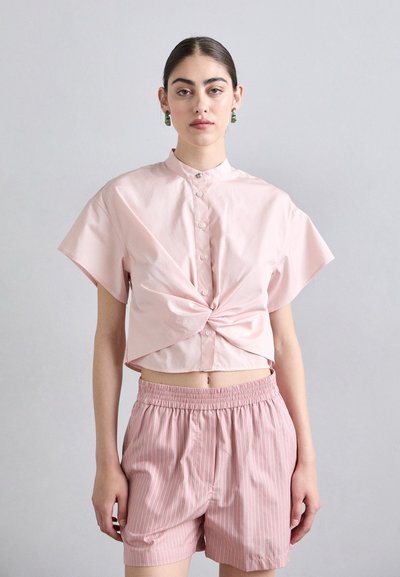 Chemisette à manches courtes, cropped, de couleur rose clair, avec un détail de nœud à l'avant et une fermeture à boutons. Associée à un short rayé rose.