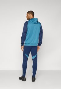 Blå og marineblå atletisk hættetrøje med hætte, parret med tætsiddende marineblå leggings med turkise accenter og diagonale mønstre.
