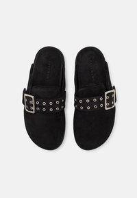 Mules en daim noir avec une large lanière munie d'œillets en argent et d'une boucle. Conception à l'arrière ouvert et doublure intérieure texturée pour plus de confort.