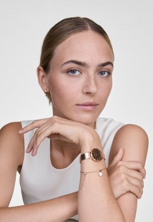 Rosegold-Netzarmbanduhr mit einem dunkelbraunen Zifferblatt. Zusätzliches zartes Armband mit einer herabhängenden weißen Perle und einem dünnen Metallelement.