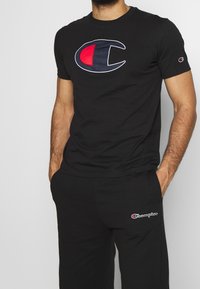Maglietta in cotone nera con ampio logo ricamato rosso e blu sul davanti; abbinata a pantaloni della tuta neri con un piccolo logo sulla coscia.