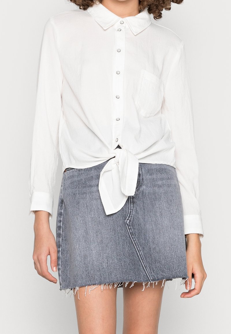 Femme portant une chemise blanche à manches longues boutonnée et nouée à la taille, ainsi qu'une mini-jupe en jean grise délavée avec un ourlet effiloché.