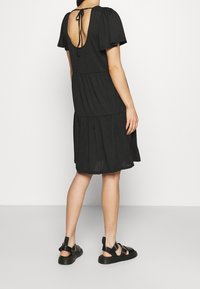 Robe noire à volants avec manches courtes bouffantes, dos ouvert avec détail noué, et longueur au-dessus du genou. Associée à des sandales noires à lanières.