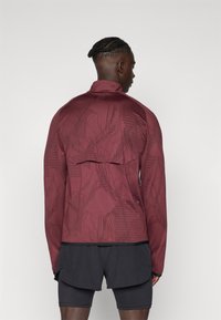 Bordo sportska jakna s geometrijskim uzorcima, slim fit, visokim ovratnikom i dugim rukavima. Nosena uz crne šorts, dizajnirana za aktivno nošenje.