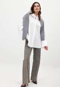 Witte button-up shirt met oversized mouwen, grijs gebreid sweater over de schouders, lichtbruine hoge taille flared jeans, donkere hakken.