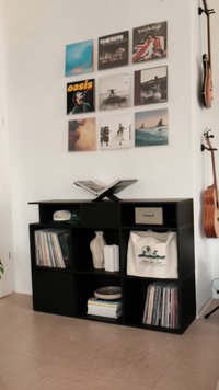 Estante de madeira preta com cubos abertos. Contém livros, discos, vasos decorativos e uma bolsa de tecido. A parede exibe discos de vinil.