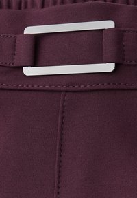 Detailní záběr maroon látkového pásku s prošitým švem a obdélníkovou stříbrnou kovovou sponou připevněnou k elastickému pásu.