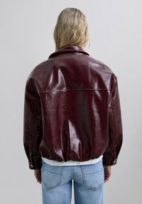 Veste en cuir bordeaux avec une finition brillante, silhouette oversize, grand col et bord élastique. Stylée avec une chemise blanche en dessous.