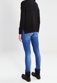 Musta neulepaita löysällä istuvuudella, yhdistettynä sinisiin skinny-farkkuihin ja nilkkureihin. Neuleessa näkyvät saumat ja tekstuuri.