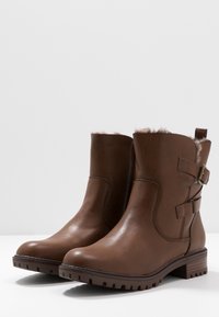 Botas de tobillo de cuero marrón con forro de piel suave, detalle de hebilla lateral, punta redondeada y suela de goma texturizada con surcos de tracción.