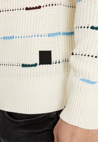 Maglione crema lavorato a maglia con strisce orizzontali multicolore in blu, verde e bordeaux. Presenta polsini a coste e un'etichetta del marchio nera.