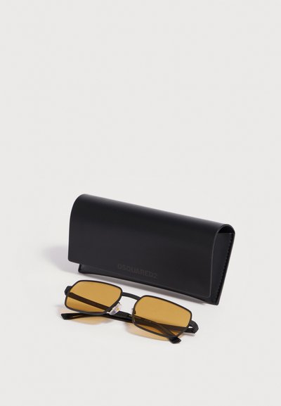 Lunettes de soleil rectangulaires avec des verres ambrés et une fine monture noire placées à côté d’un étui DSQUARED2 noir sur une surface blanche.